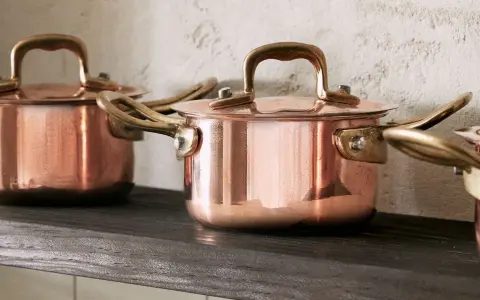Drei glänzende Kupfertopf-Sets mit Messinggriffen stehen auf einem dunklen Regal. Sie warten darauf, mit einem speziellen Mittel zum Kupfer reinigen wieder zum Strahlen gebracht zu werden.