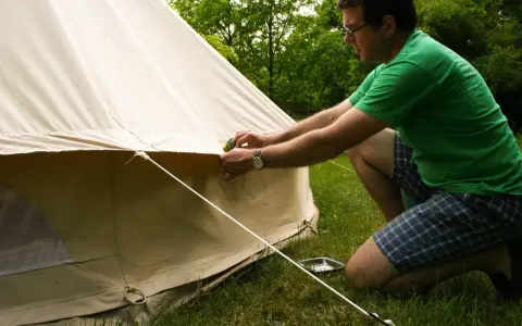 Mann reinigt mit einem Schwamm das beige Zelt. Er kniet auf dem Gras neben dem Zelt.