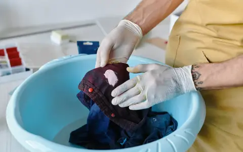 Hände mit Handschuhen behandeln einen Fleck auf einem dunkelblauen Hemd in einer blauen Schüssel. Tipps zur Entfernung von Fettflecken aus Polyester.
