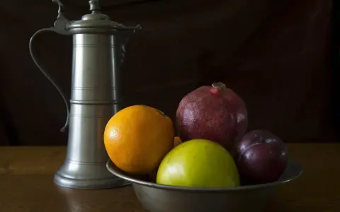 Ein Zinnkrug steht neben einer Schale mit Früchten: Orange, Apfel, Granatapfel und Pflaume. Zeit für zinn reinigen.