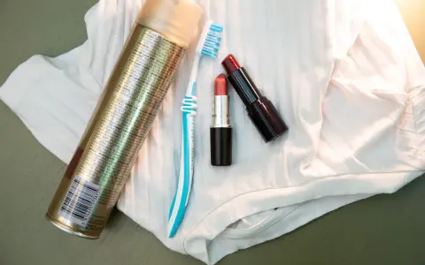 Haarspray, Zahnbürste und Lippenstifte liegen auf weißem Stoff. Ein Tipp, wie man Lippenstift aus Kleidung entfernen kann.