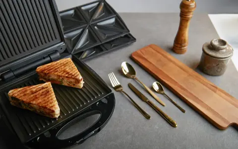 Zwei goldbraune Sandwiches liegen im geöffneten Sandwichmaker. Daneben liegen Besteck, ein Holzbrett und Gewürzmühlen. Zeit, den Sandwichmaker zu reinigen.