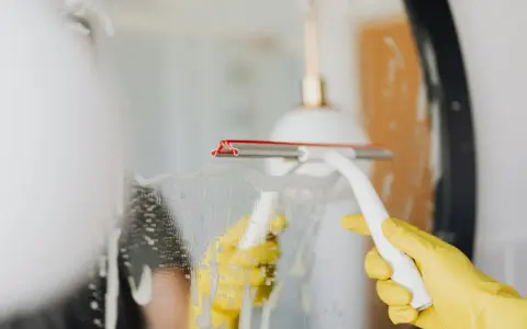 Gelbe Handschuhe und ein Wischer reinigen einen beschlagenen Spiegel.
