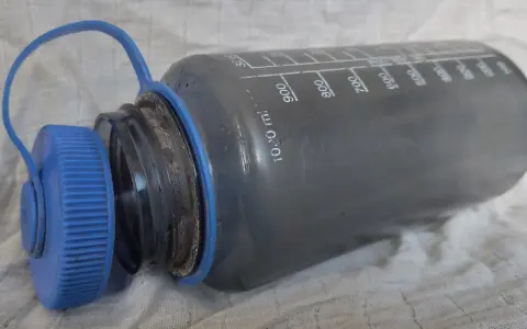 Graue Trinkflasche mit blauer Kappe, die eine gründliche Reinigung benötigt. Messskala bis 1000 ml sichtbar.