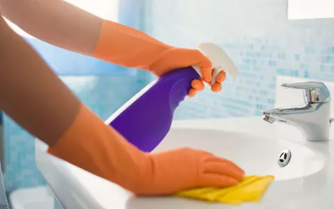 Hände in orangen Handschuhen sprühen Reinigungsmittel auf ein Waschbecken und wischen mit einem gelben Putzlappen.
