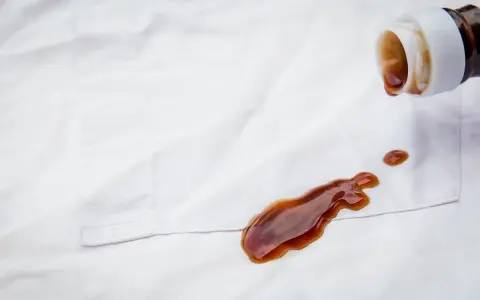 Braune Flüssigkeit tropft aus einer Flasche auf weiße Kleidung. Tipps zur **balsamico flecken entfernen** sind hier gefragt.