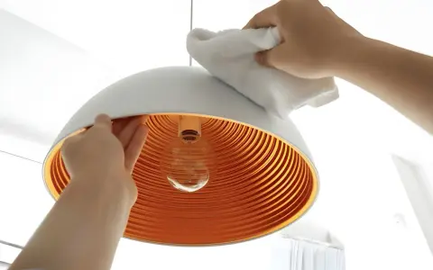 Hände reinigen einen modernen Lampenschirm mit einem weißen Tuch. Das Innere des Schirms ist orange und geriffelt.