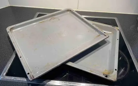Zwei alte, graue Backbleche mit Gebrauchsspuren und Verfärbungen liegen übereinander auf einem Cerankochfeld. Zeit, das Backblech reinigen!