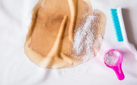 Brauner Fleck auf weißem Stoff, dane Waschpulver in einem rosa Löffel und eine blaue Bürste. So lässt sich **Haarfarbe aus Kleidung entfernen**.