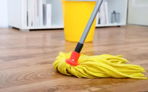 Gelber Wischmopp auf Laminatboden, bereit für die Reinigung mit Hausmitteln. Ein gelber Eimer steht im Hintergrund.