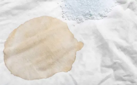 Ein brauner Fleck auf weißem Stoff, dane ein Haufen Waschpulver. So lassen sich hartnäckige teeflecken entfernen.
