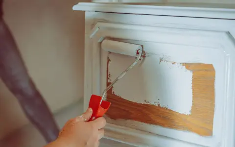 Eine Hand mit einem Farbroller trägt weiße Farbe auf ein Holzmöbel auf. Das Holz wird vorbereitet, um es zu entfetten und neu zu streichen.