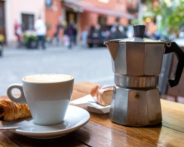 Ein Espresso-Kocher steht neben einer Tasse Cappuccino und einem Croissant auf einem Holztisch. Nach dem Genuss ist das espressokocher reinigen angesagt.