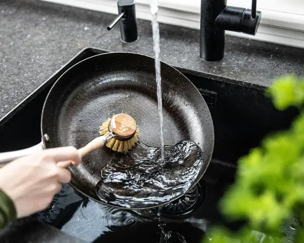 Eine Hand schrubbt mit einer Bürste eine Gusseisenpfanne unter fließendem Wasser. So gelingt das Gusseisenpfanne reinigen mühelos.