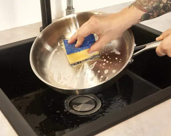 Hand reinigt mit einem Schwamm den schmutzigen Pfannenboden in der Spüle.