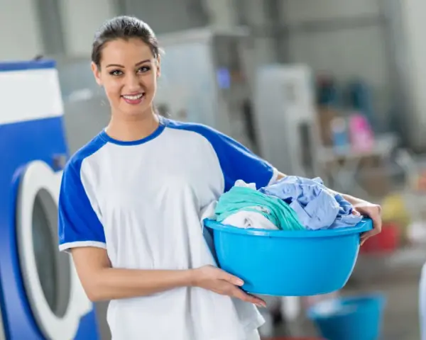 Lächelnde Frau mit Wäschekorb voller bunter Kleidung vor einer Waschmaschine. Sie weiß, wie man richtig waschen.