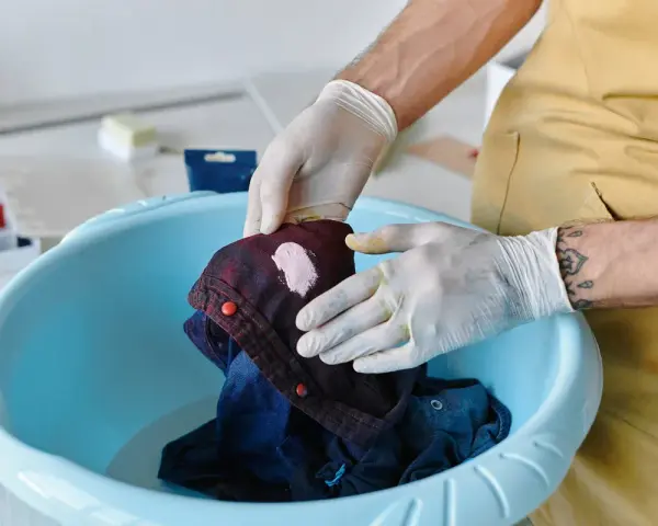 Hände mit Handschuhen behandeln einen Fleck auf einem dunkelblauen Hemd in einer blauen Schüssel. Tipps zur Entfernung von Fettflecken aus Polyester.