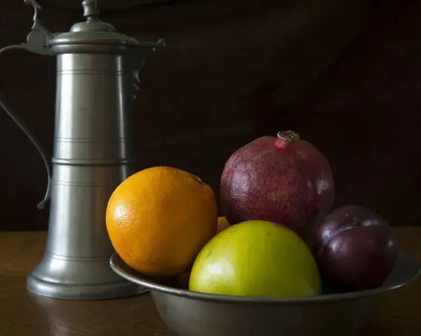 Ein Zinnkrug steht neben einer Schale mit Früchten: Orange, Apfel, Granatapfel und Pflaume. Zeit für zinn reinigen.