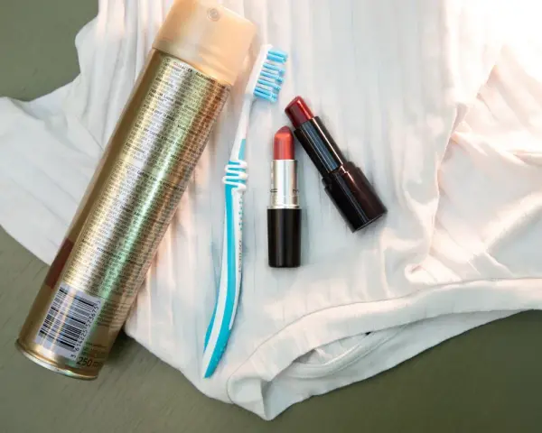 Haarspray, Zahnbürste und Lippenstifte liegen auf weißem Stoff. Ein Tipp, wie man Lippenstift aus Kleidung entfernen kann.