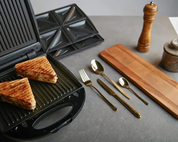 Zwei goldbraune Sandwiches liegen im geöffneten Sandwichmaker. Daneben liegen Besteck, ein Holzbrett und Gewürzmühlen. Zeit, den Sandwichmaker zu reinigen.