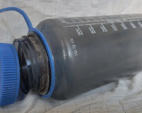 Graue Trinkflasche mit blauer Kappe, die eine gründliche Reinigung benötigt. Messskala bis 1000 ml sichtbar.