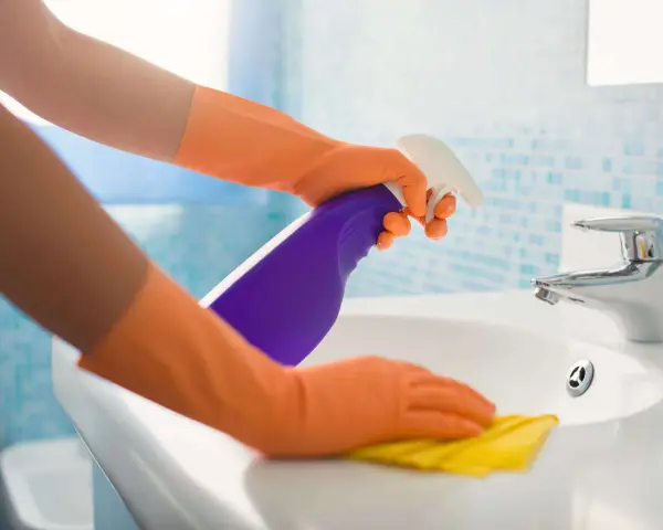 Hände in orangen Handschuhen sprühen Reinigungsmittel auf ein Waschbecken und wischen mit einem gelben Putzlappen.