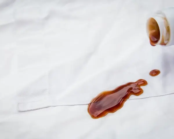 Braune Flüssigkeit tropft aus einer Flasche auf weiße Kleidung. Tipps zur **balsamico flecken entfernen** sind hier gefragt.