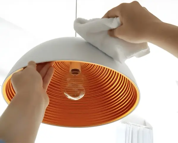 Hände reinigen einen modernen Lampenschirm mit einem weißen Tuch. Das Innere des Schirms ist orange und geriffelt.