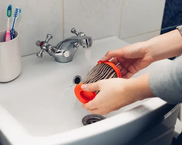 Hände reinigen einen staubigen Staubsaugerfilter unter fließendem Wasser im Waschbecken.