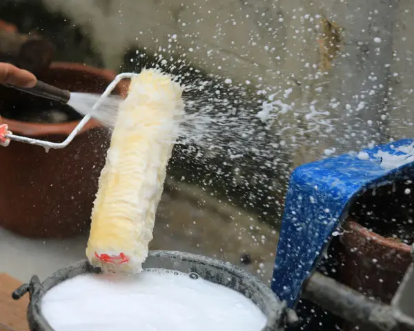 Eine Hand spritzt Wasser auf eine Malerrolle, um sie in einem Eimer mit Seifenwasser zu reinigen.