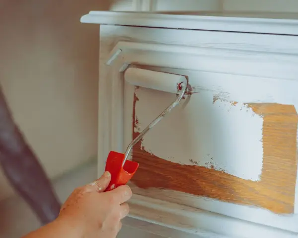 Eine Hand mit einem Farbroller trägt weiße Farbe auf ein Holzmöbel auf. Das Holz wird vorbereitet, um es zu entfetten und neu zu streichen.