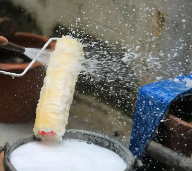 Eine Hand spritzt Wasser auf eine Malerrolle, um sie in einem Eimer mit Seifenwasser zu reinigen.