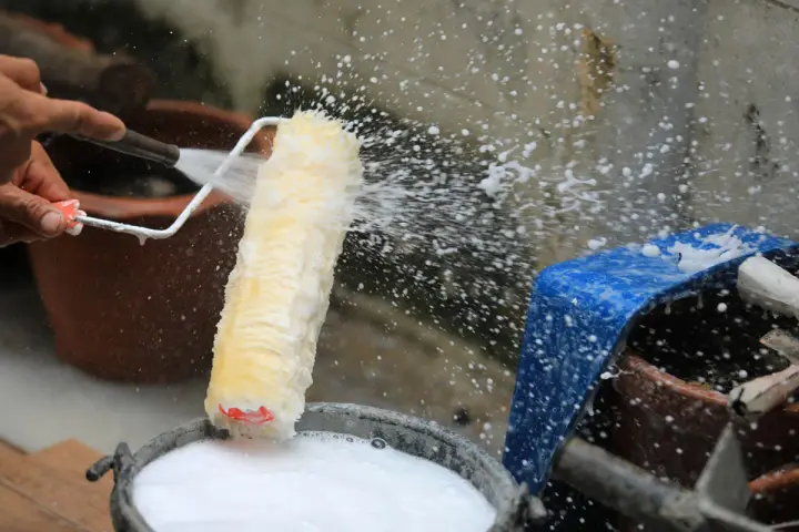 Eine Hand spritzt Wasser auf eine Malerrolle, um sie in einem Eimer mit Seifenwasser zu reinigen.