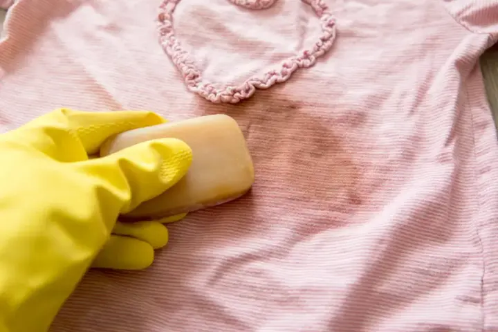Gelbe Handschuhe halten Gallseife, um einen Fleck auf einem rosafarbenen gestreiften Shirt zu behandeln.