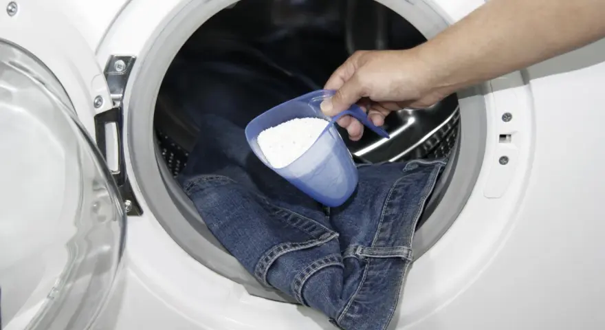 Eine Hand gibt Waschpulver in eine Waschmaschine, um Kettenfett aus Jeans zu entfernen.
