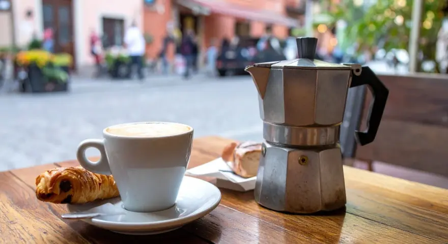 Ein Espresso-Kocher steht neben einer Tasse Cappuccino und einem Croissant auf einem Holztisch. Nach dem Genuss ist das espressokocher reinigen angesagt.