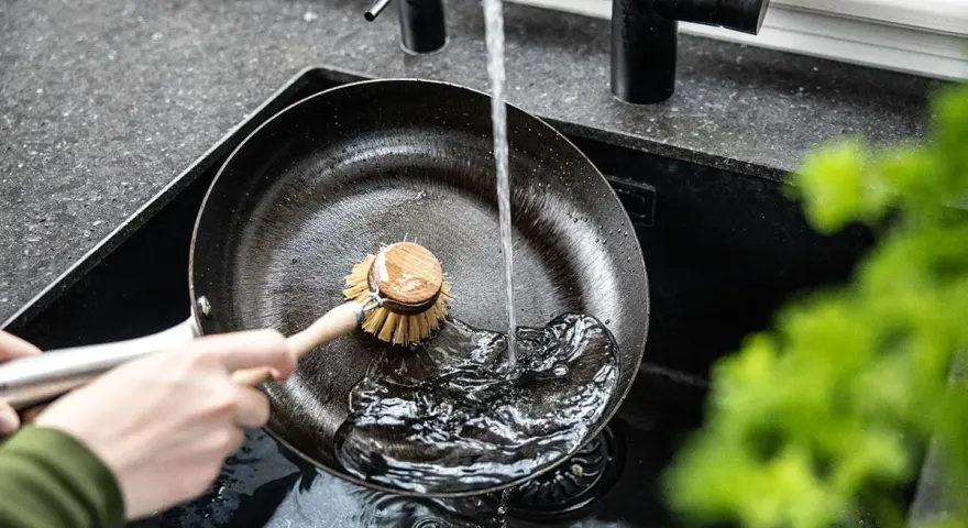 Eine Hand schrubbt mit einer Bürste eine Gusseisenpfanne unter fließendem Wasser. So gelingt das Gusseisenpfanne reinigen mühelos.