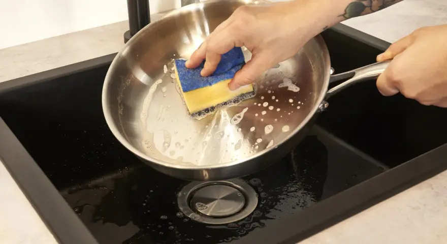Hand reinigt mit einem Schwamm den schmutzigen Pfannenboden in der Spüle.