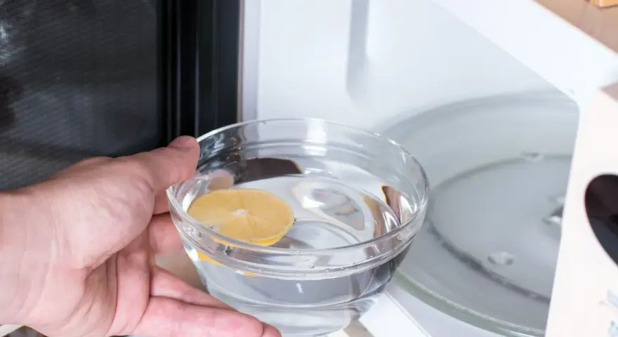 Hand placing a bowl of water with lemon slice into a microwave for cleaning.