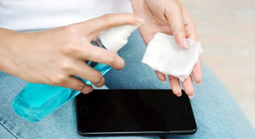 Hände desinfizieren: Eine Person sprüht Desinfektionsmittel auf ein Wattepad, um das Handy zu reinigen.