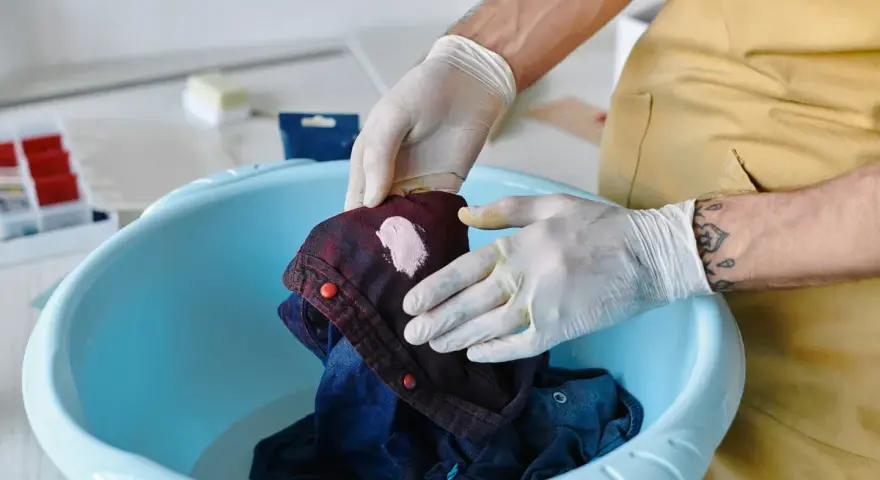 Hände mit Handschuhen behandeln einen Fleck auf einem dunkelblauen Hemd in einer blauen Schüssel. Tipps zur Entfernung von Fettflecken aus Polyester.