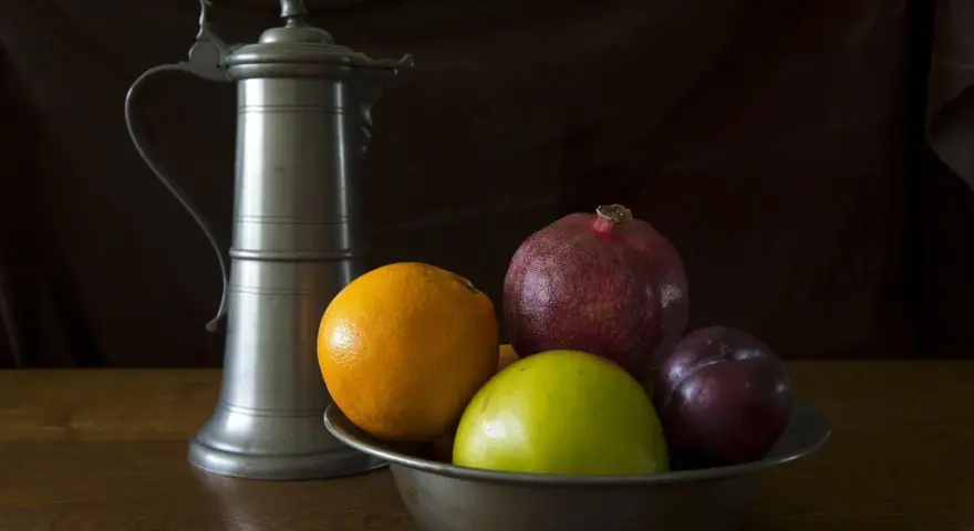 Ein Zinnkrug steht neben einer Schale mit Früchten: Orange, Apfel, Granatapfel und Pflaume. Zeit für zinn reinigen.