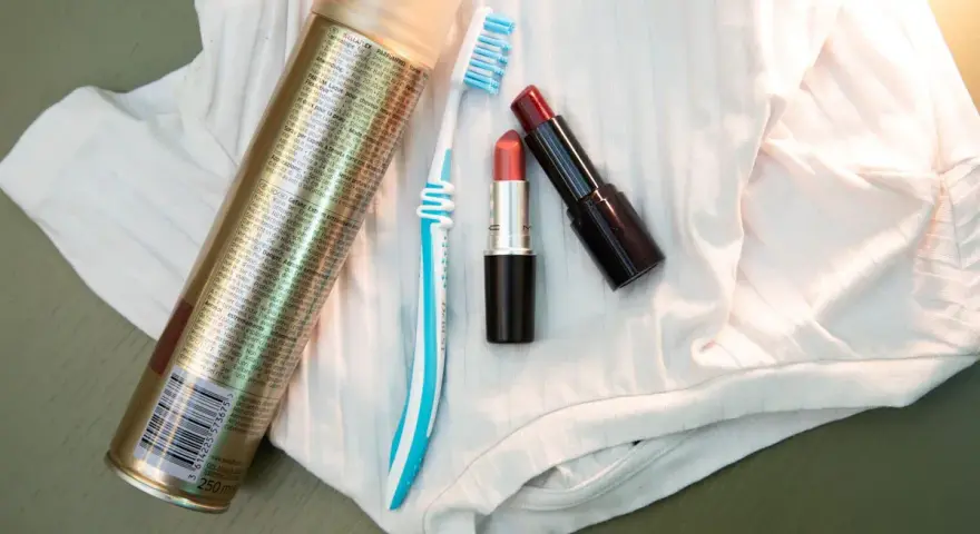 Haarspray, Zahnbürste und Lippenstifte liegen auf weißem Stoff. Ein Tipp, wie man Lippenstift aus Kleidung entfernen kann.