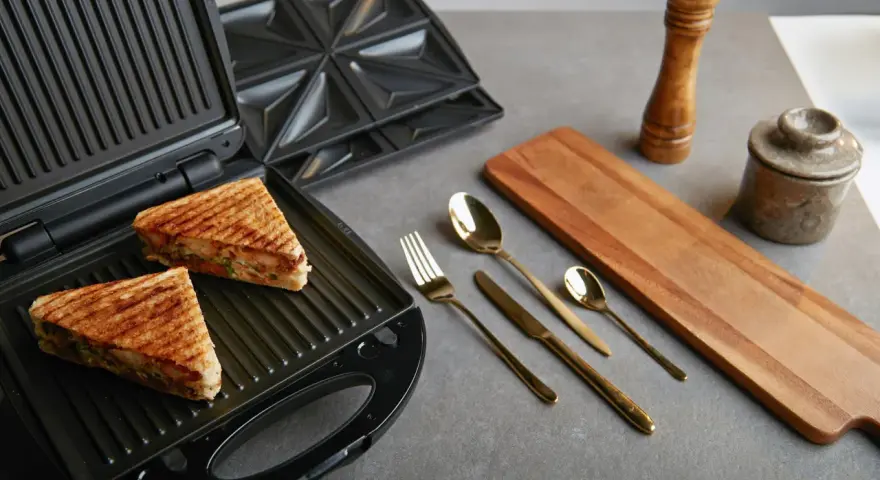 Zwei goldbraune Sandwiches liegen im geöffneten Sandwichmaker. Daneben liegen Besteck, ein Holzbrett und Gewürzmühlen. Zeit, den Sandwichmaker zu reinigen.