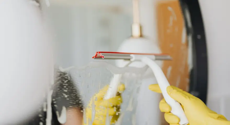 Gelbe Handschuhe und ein Wischer reinigen einen beschlagenen Spiegel.