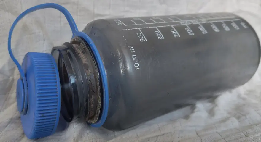 Graue Trinkflasche mit blauer Kappe, die eine gründliche Reinigung benötigt. Messskala bis 1000 ml sichtbar.