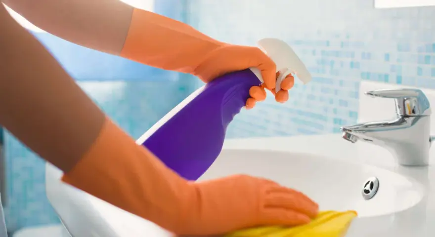 Hände in orangen Handschuhen sprühen Reinigungsmittel auf ein Waschbecken und wischen mit einem gelben Putzlappen.