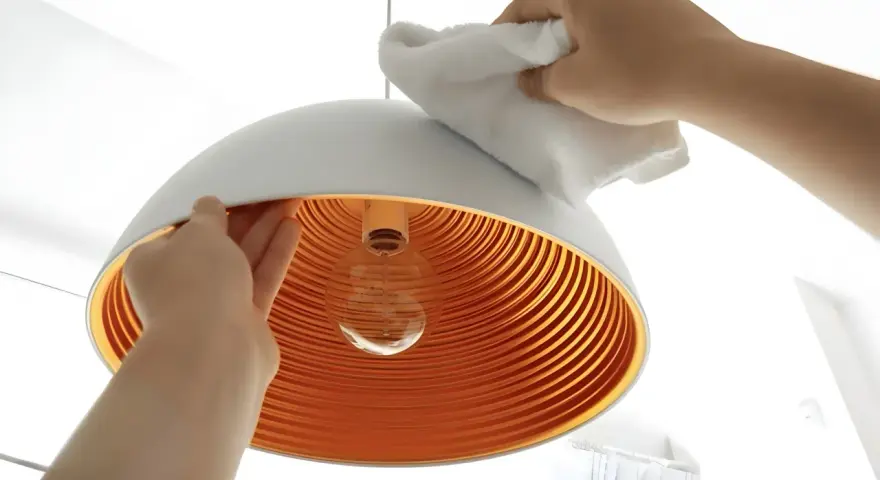 Hände reinigen einen modernen Lampenschirm mit einem weißen Tuch. Das Innere des Schirms ist orange und geriffelt.