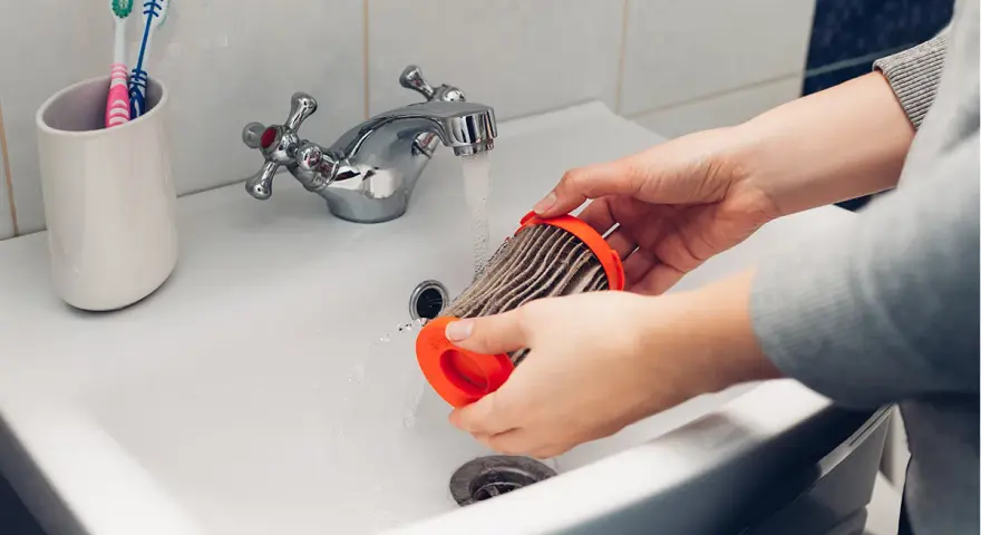 Hände reinigen einen staubigen Staubsaugerfilter unter fließendem Wasser im Waschbecken.