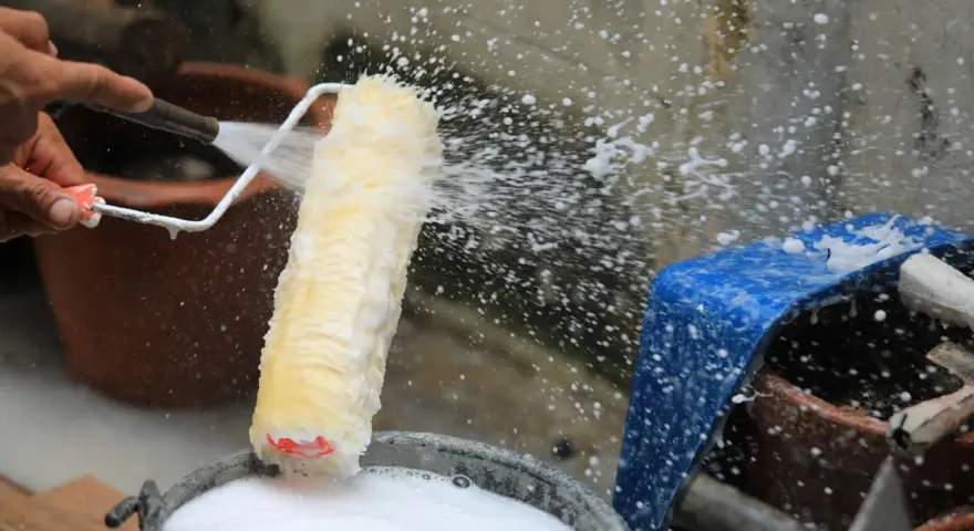 Eine Hand spritzt Wasser auf eine Malerrolle, um sie in einem Eimer mit Seifenwasser zu reinigen.